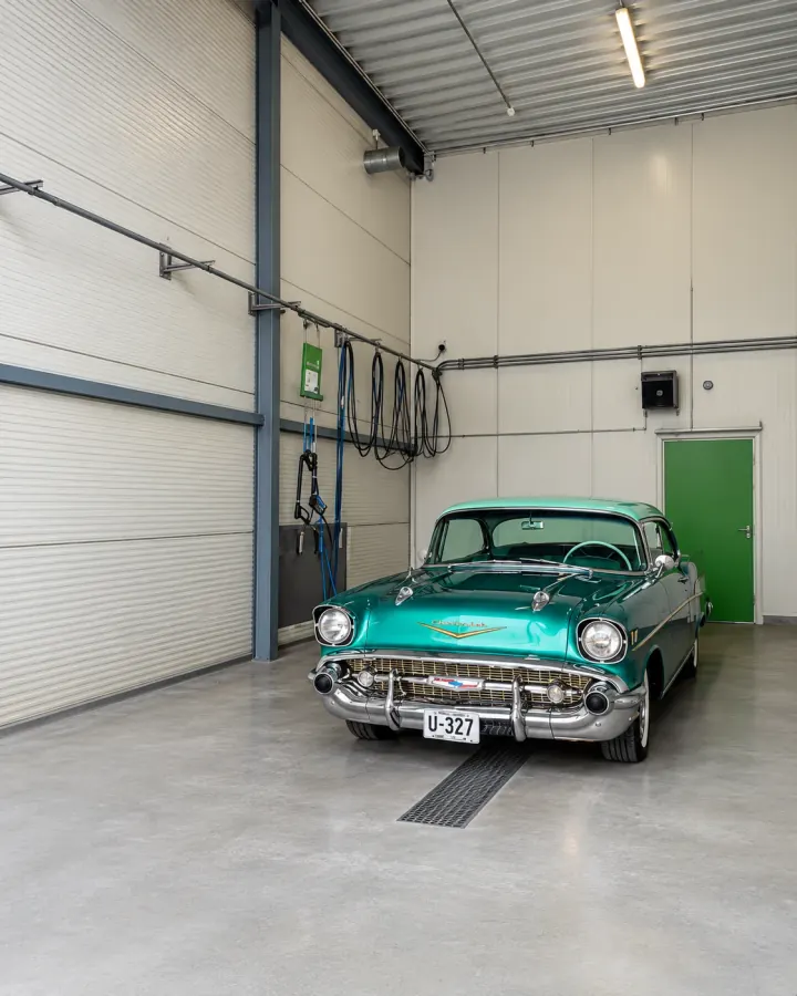 Innsiden av vaskehallen med lyse vegger, slanger på stativ, sluk i gulvet og en grønn Chevrolet fra 1950-tallet